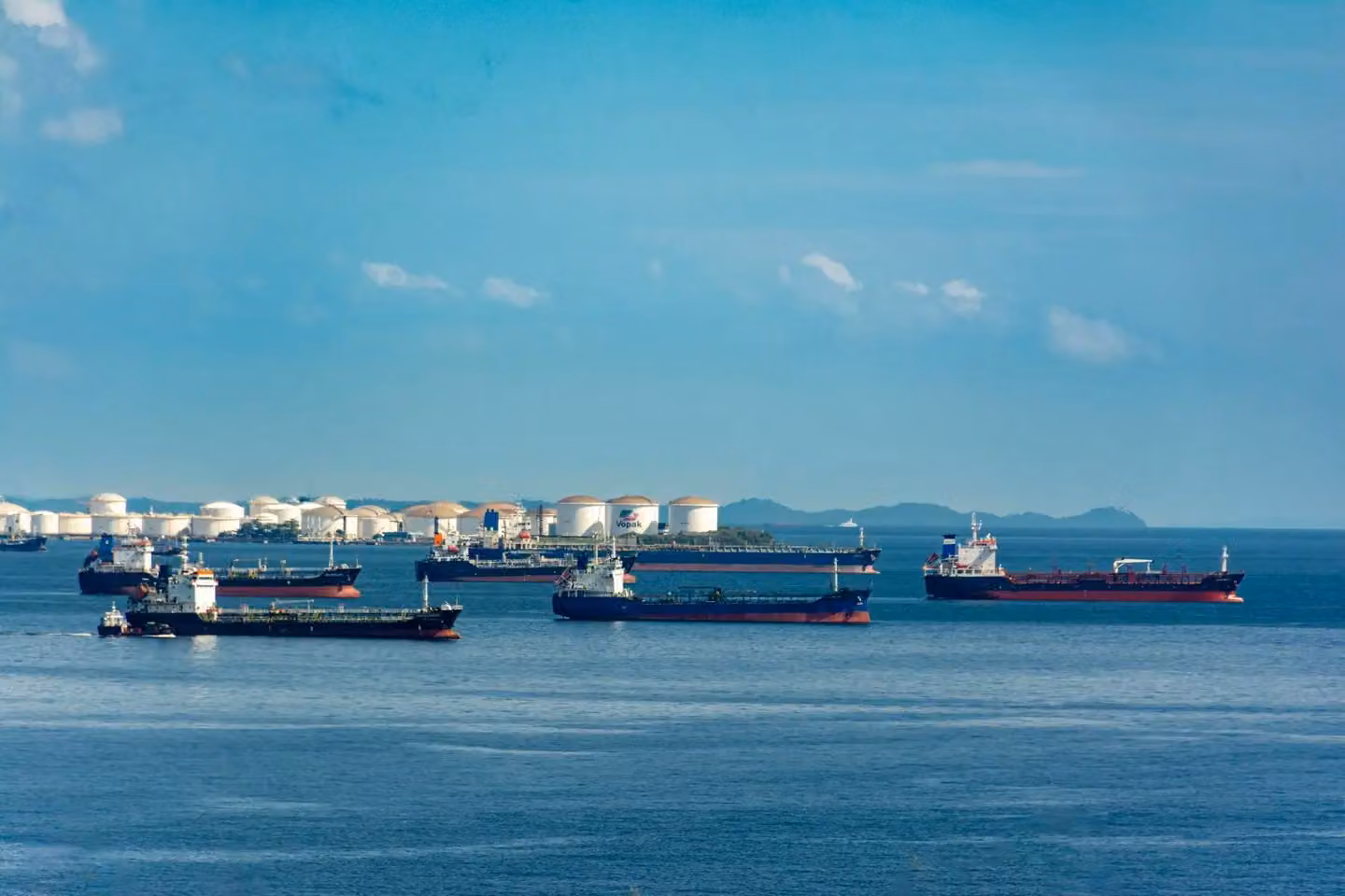 A fleet of cargo ships docked near oil storage tanks along a serene coastline with a clear blue sky above.