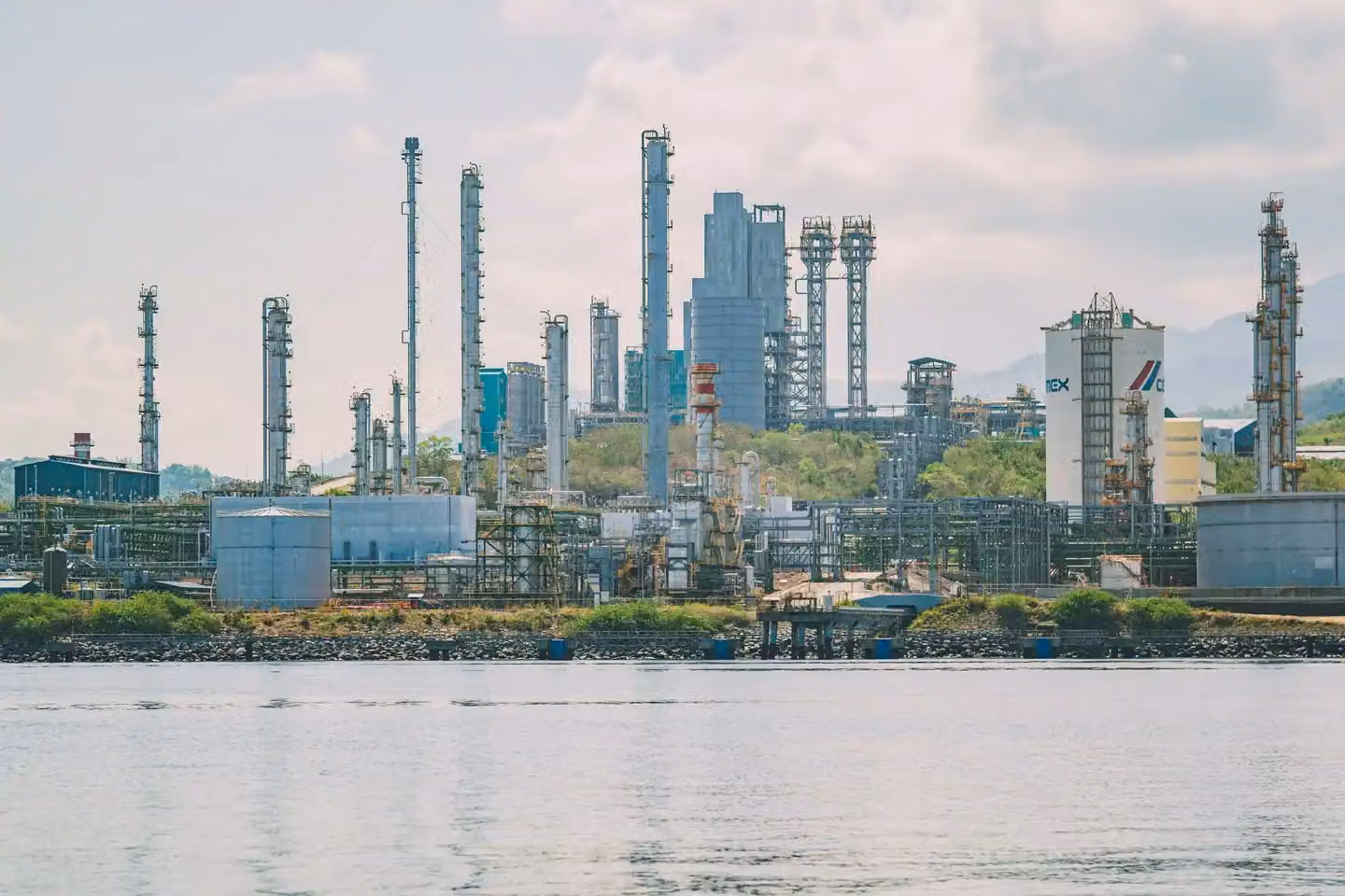 A large industrial refinery complex with tall chimneys by the waterfront under a clear sky.