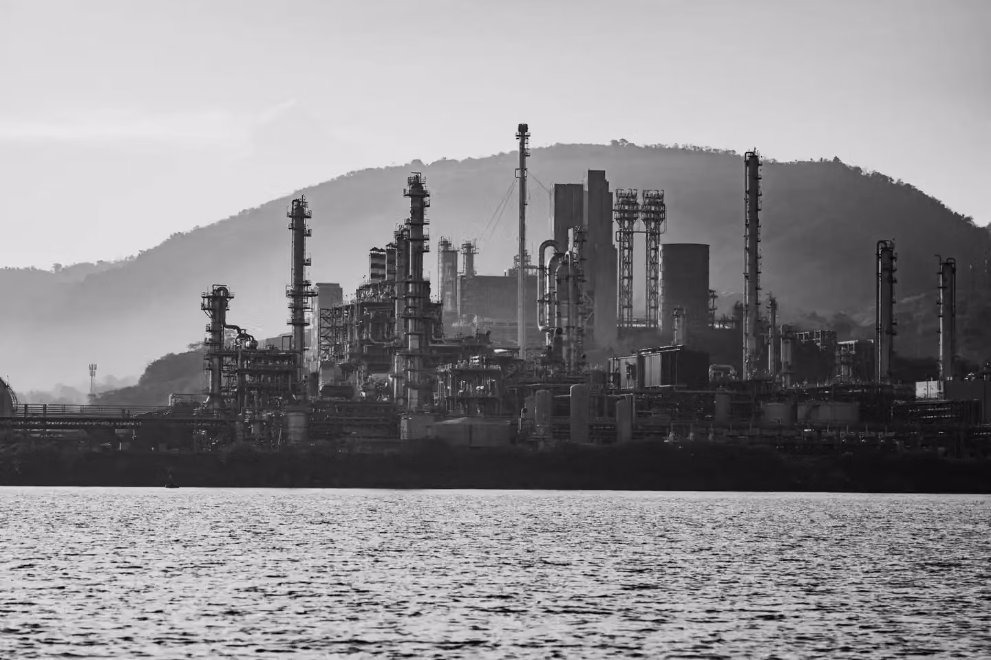 Black and white image of an industrial refinery set against a mountainous backdrop with water in the foreground.