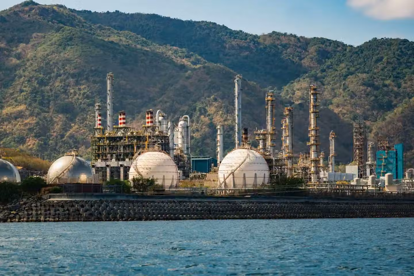 Industrial refinery with spherical tanks by the coastline against a backdrop of mountains.