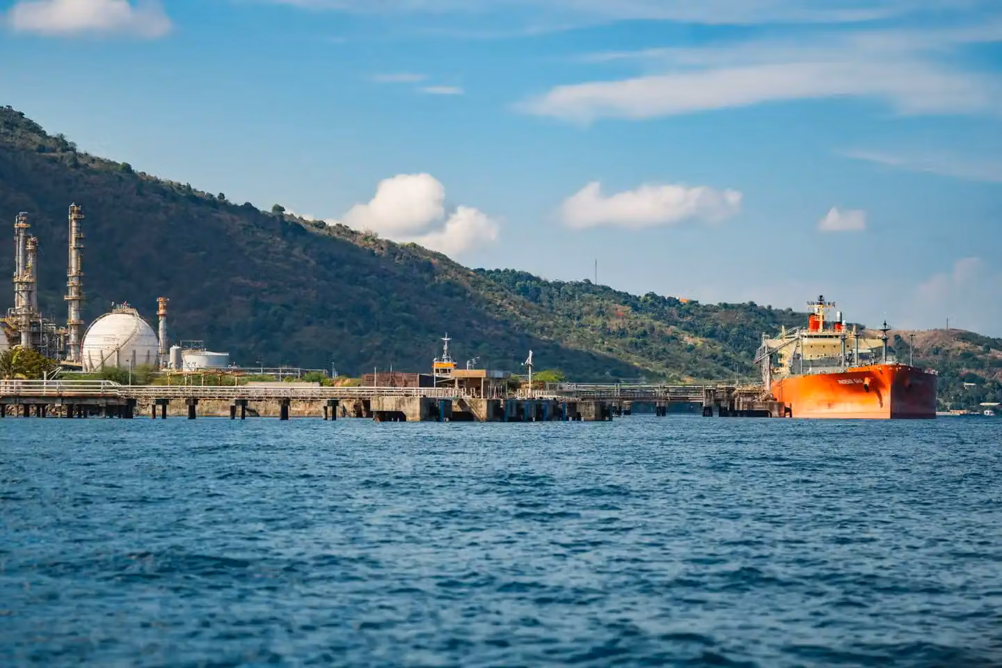 A large oil tanker docked at an industrial port with refinery and mountainous backdrop.