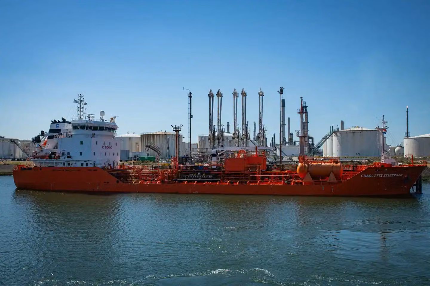 Large red tanker ship docked at an industrial port with storage tanks and chimneys under a clear blue sky.